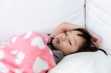 3 yaşındaki Asyalı bir kız, beyaz yatakta, pembe battaniyenin altında, Asyalı bir çocuğa ve sağlık hizmetleri konseptine karşı mışıl mışıl uyuyor..