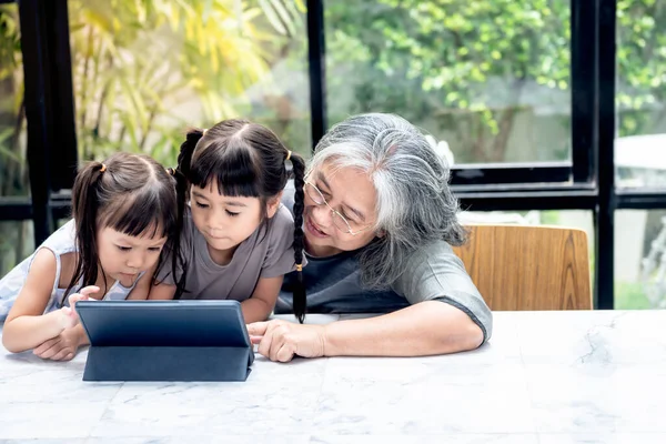 Asian family, Elderly grandmather and 2 daughters looking at tablet on marble table in the house, with blur background, to Asian family and children education online concept.