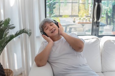 Asyalı şişman bir adam, beyaz koltukta oturup dinleniyor ve kulaklık takarak müzik dinliyor..