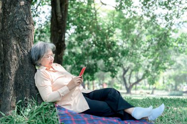 Asyalı yaşlı bir kadın, bahçedeki yeşil çimlerin üzerinde oturan ve rahatlatıcı bir cep telefonu kullanarak emeklilik yaşına ve doğa kavramına mutlulukla bakıyor..