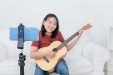 Kanepede oturan Asyalı bir kadının bulanık görüntüleri internet kanallarından cep telefonuyla gitar çalarken çevrimiçi müzik dersleri alırken görülüyor. Bu resim cep telefonuna odaklanıyor.