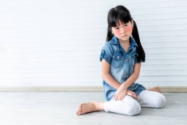 Asyalı bir kızın portre resimleri 5 yaşında, gözlük takıyor ve beyaz duvar arkaplanlı yerde oturuyor..