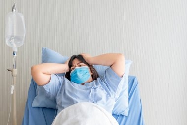 Asyalı şişman kadın hastalar cerrahi maske takıyor. Bir hastanın yatağında yatıyorlar. Şiddetli baş ağrıları çekiyorlar..