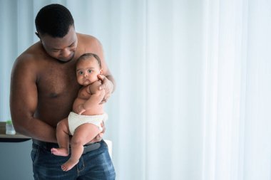 Afrikalı bir baba kızını kucağında tutuyor. 3 aylık yeni doğmuş bir bebek. Beyaz bir yatak odasında güvende ve mutlu.