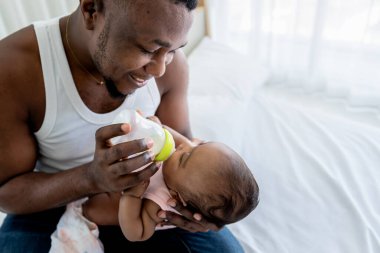Afrikalı baba yatakta oturuyor ve sütü şişeden yeni doğmuş bebeğine, 3 aylık kızına, ailesine ve yeni doğmuş bebek konseptine yediriyor..