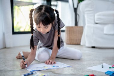 Yerde yatan 5 yaşındaki Asyalı bir kızın bulanık görüntüleri, renkli kalemler, çocuklar için konsept ve çevrimiçi eğitim kullanarak sanat yapıyorlar..