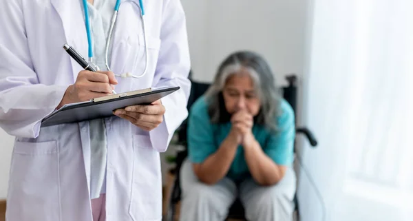The doctor writing report for record the patient\'s condition, with blur background of Asian female elderly patient sitting in a wheelchair and have stress, to elderly and health care concept.