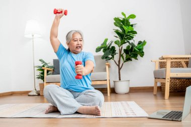 Asian elderly woman doing exercise at home by stretching the arm muscles and using a dumbbell as an exercise aid, to people retirement age and health care concept.