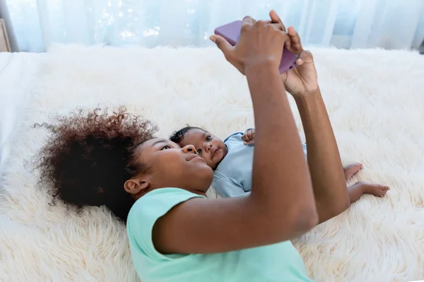 African-American family, Mother and 1 month old baby newborn son lying on white bed and looking at mobile phone, while mom takes a selfie, with smile and happy, to relationship family and infant newborn concept.