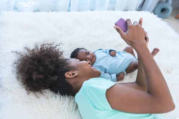 African American family, Mother and 1 month old baby newborn son lying on white bed and looking at mobile phone, while mom takes a selfie, with smile and happy, to family and infant newborn concept.