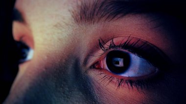 Extreme closeup of caucasian woman surfing internet at night, using a device before sleep with insomnia or sleeplessness. Screen with social network reflecting in her eyes. Internet addiction concept