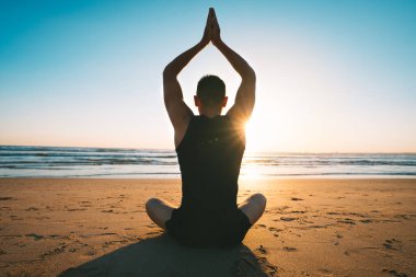 Kumda oturup yoga yapan ya da gün batımında okyanus manzaralı sahilde meditasyon yapan bir adam.