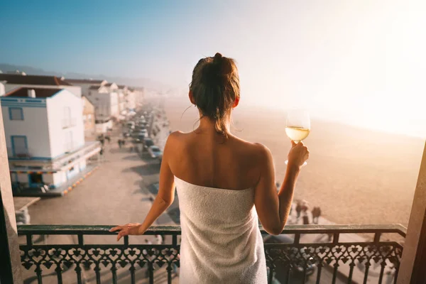 Donna avvolta in asciugamano in piedi sul balcone e con un bicchiere di vino bianco in un appartamento di lusso con vista sull'oceano. Donna in vacanza rilassante e godersi la vita — Foto Stock