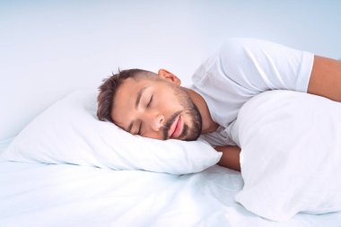 Beyaz yataklı yatakta uyuyan yakışıklı bir adam. Adam yastığa uzanıyor ve sağlıklı bir uykudan zevk alıyor.