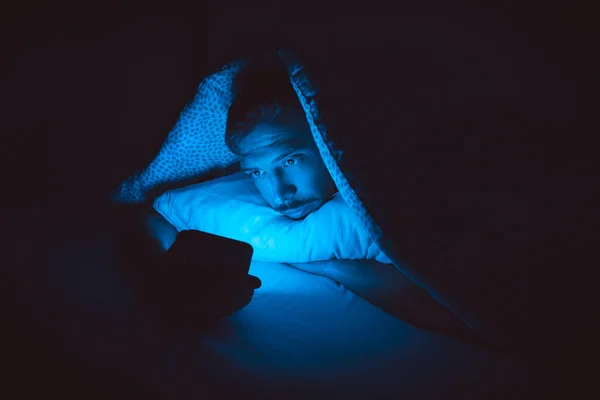 Młody człowiek leży w łóżku i używa smartfona przed snem. Uzależnienie od mediów społecznościowych i bezsenność — Zdjęcie stockowe