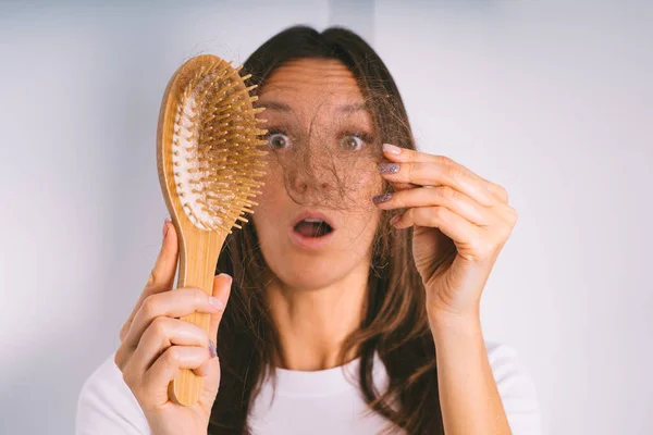 Jovem mulher chocada por causa do problema de perda de cabelo. Mulher segurando escova de cabelo e mostrando cabelo danificado. Cabelo ruim caindo — Fotografia de Stock