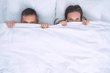 Çekici bir çift yatakta uzanıyor ve yüzünü battaniyeyle örtüyor ya da beyaz battaniyenin altına saklanıyor. İlişki ve aşk kavramı. Boşluğu kopyala