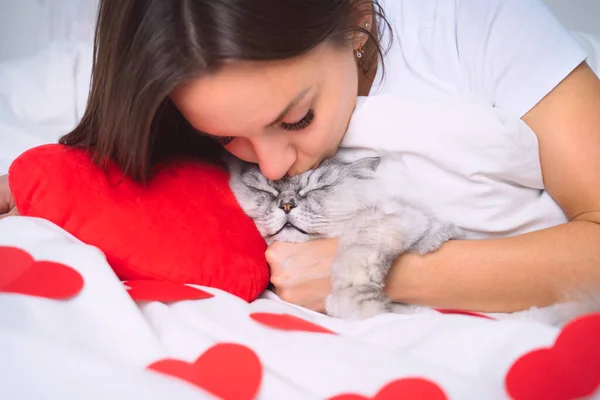 Женщина лежит на белой кровати с красными сердцами и целует свою милую кошку. Кот спит с владельцем. Домашние животные доверие и любовь — стоковое фото