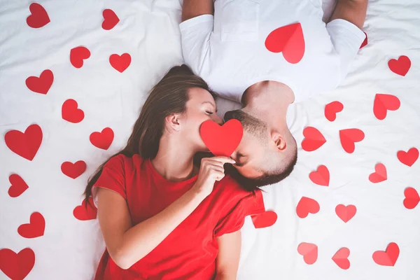 Horní pohled na krásný pár v lásce ležící na bílé posteli s červenými srdci a líbání. Koncept lásky a vztahů. Valentines day concept — Stock fotografie