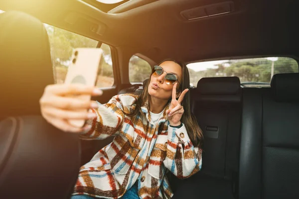 Stijlvolle jonge vrouw zit op de achterbank in de auto en maakt selfie met smartphone. Vrouw neemt foto met telefoon terwijl ze in de taxi zit — Stockfoto