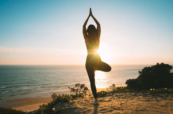 Woman practicing yoga and meditates on the mountain with ocean view at sunset. Young woman standing in vrksasana exercise, tree pose outdoor in the nature — Foto Stock
