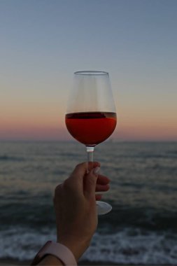 Bir bardak kırmızı şarap ve deniz kenarında pembe bir gün batımı.