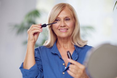 Mutlu bir son sınıf öğrencisi evde oturup kirpiklerine rimel sürüyor. Makyaj yapan yaşlı bir kadın.