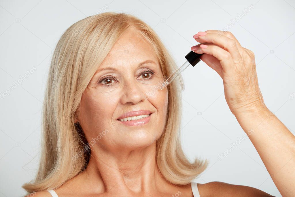 A happy blond senior lady trying out new serum. Beauty photography