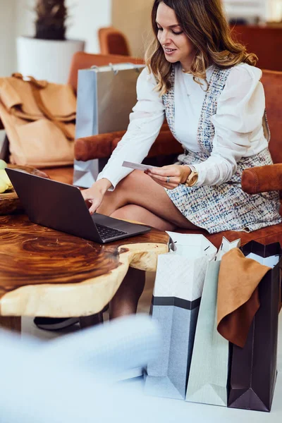 一个穿着连衣裙的快乐女人正坐在咖啡店网上买新衣服 她的腿旁边有购物袋 — 图库照片