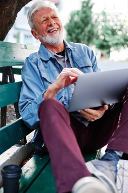 Mutlu bir son sınıf öğrencisi elinde dizüstü bilgisayarla parkta oturuyor ve dizüstü bilgisayar kullanıyor. Yaşlılar için geri çekiliş.