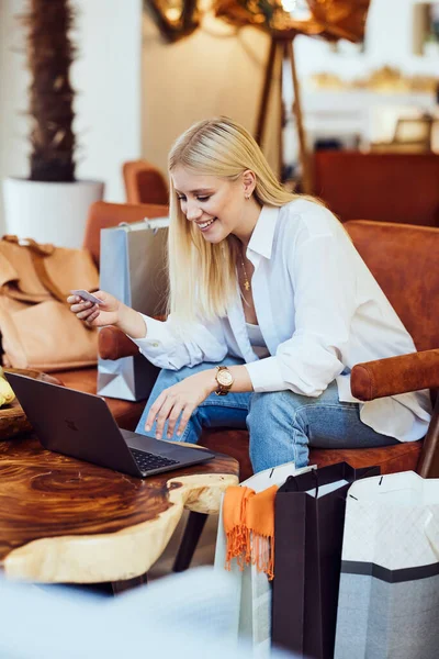 一位年轻的金发美女坐在咖啡店里 在笔记本电脑上输入信用卡号码 她的腿旁边有购物袋 女人上网买东西 — 图库照片