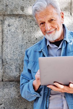 Mutlu bir son sınıf öğrencisi tuğla duvarın önünde dizüstü bilgisayarda yazı yazıyor..