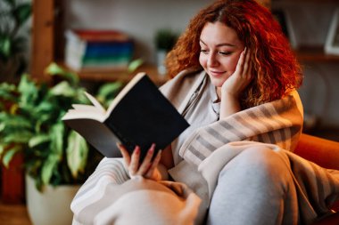 Oturma odasında battaniyeye sarılmış güzel bir kızıl kız sabahları ilginç bir kitap okuyor..