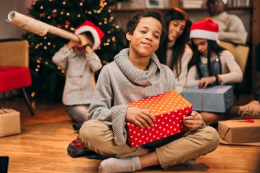 Mutlu bir Afro-Amerikalı çocuk elinde Noel hediyesiyle evde yerde oturur ve kameraya bakar. Bulanık arka planda ailesi hediyeleri açıyor..