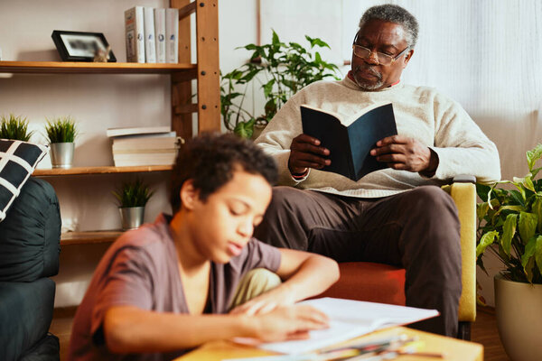 An African grandad sits at home, reads a book, and keeps company to a grandson. A grandchild is doing his math homework. Generations and age difference