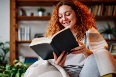 Evde kızıl saçlı bir kitap okuyor. Mutlu bir kızıl kız battaniyeyle kaplı bir koltukta oturur ve evinde ilginç bir kitap okur..