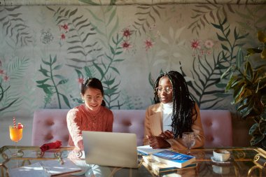 İki zeki üniversiteli kız bir kahve dükkanında ellerinde kitaplarla oturuyorlar ve online ders için dizüstü bilgisayar kullanıyorlar. Kızlar bir kahve dükkanında eğitim görüyorlar..