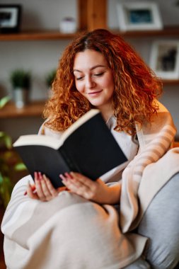 Evde kızıl saçlı bir kitap okuyor. Mutlu bir kızıl kız battaniyeyle kaplı bir koltukta oturur ve evinde ilginç bir kitap okur..