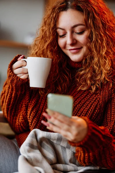 곱슬머리의 귀여운 소녀가 아침에 의자에 전화로 메시지를 보내고 커피를 마시고 — 스톡 사진