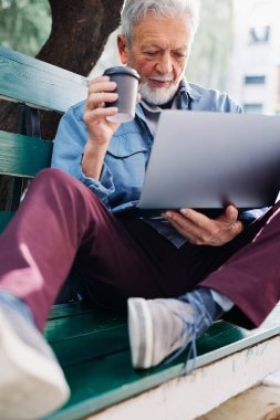 A senior student sits in a park with a laptop in his hands and using a laptop while drinking coffee. Retraining for seniors.