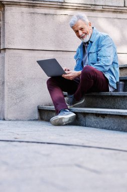 Üniversite binasının önündeki merdivenlerde elinde dizüstü bilgisayarla oturan mutlu bir son sınıf öğrencisi ders çalışıyor. Geri çekilen konsept.