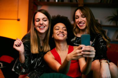 Üç çok kültürlü bayan arkadaş akşamları oturma odasında oturup ev partisinde selfie çekiyorlar..
