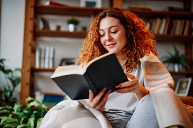 Oturma odasında battaniyeye sarılmış güzel bir kızıl kız sabahları ilginç bir kitap okuyor..