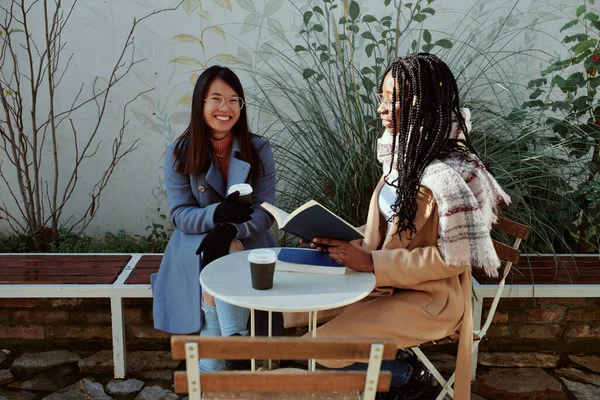 Dwie Wielokulturowe Dziewczyny Siedzące Przed Kawiarnią Pijące Kawę Czytające Książki — Zdjęcie stockowe