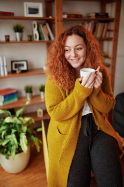 Mutlu genç bir kızıl kız sabah oturma odasında duruyor ve sabah kahvesinin tadını çıkarıyor..