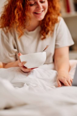 Kızıl saçlı bir kız yatakta kahvaltı yapıyor. Kızıl bir kız yatak odasında yatağında oturmuş mısır gevreği yiyor ve internette dizüstü bilgisayarla sörf yapıyor..