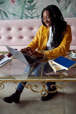 Öğrenci değişiminde olan mutlu bir Afrikalı kız öğrenci bir kafede oturuyor ve dizüstü bilgisayarında ödev yazıyor. Öğrenci değişimi.