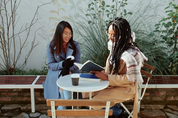 Dwie Wielokulturowe Dziewczyny Siedzące Przed Kawiarnią Pijące Kawę Czytające Książki — Zdjęcie stockowe