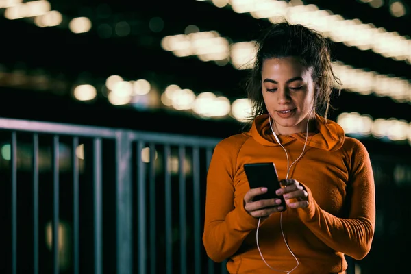 A happy female night runner is using the phone for music on a city street.