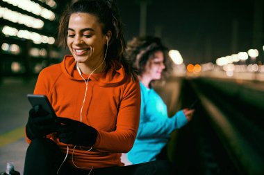 Geceleri gülümseyen, zinde bir sporcu kadın dışarıda oturuyor, dinleniyor ve telefon kullanıyor..